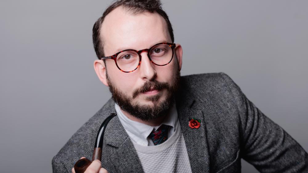 Headshot of Ross Mittiga, holding a pipe in his right hand, against a grey background