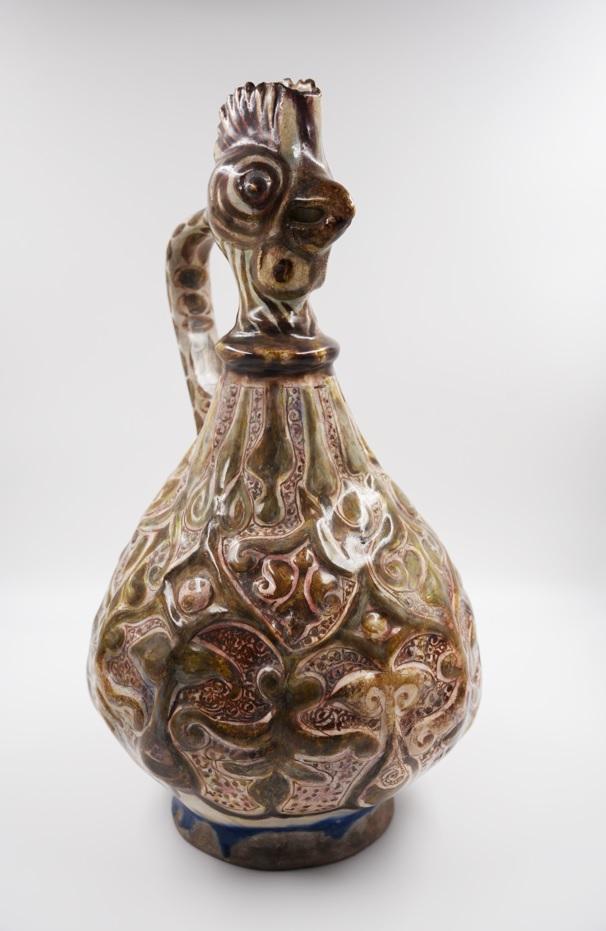 A cockerel shaped jug sits on a white background. The jug has leaf like patterns all over with several colours, including dark red, dark and light brown, with a deep blue base. 