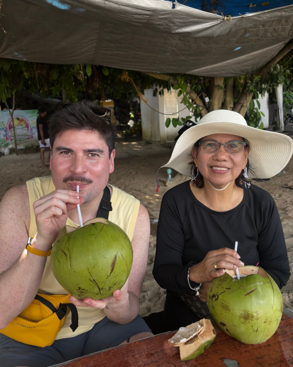 An adult son and his mum smile at the camera wearing from straws on a beach