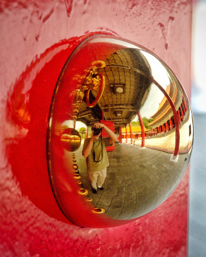Reflection in a gold doorknob on a red door captures a person taking a photo, with an ancient temple featuring ornate columns and curved roofs in the background.
