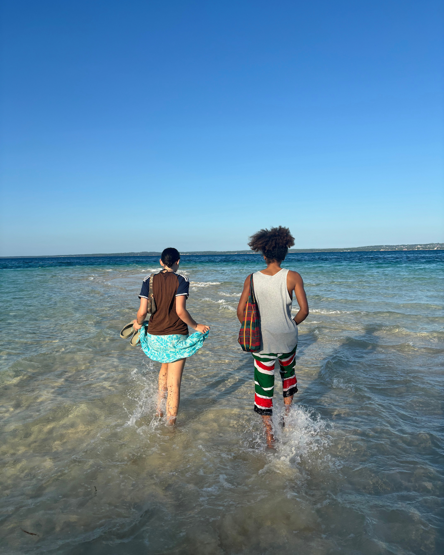 Two people walk barefoot in shallow ocean water under a clear blue sky. 