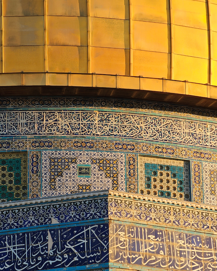 Close-up of a building's golden dome, reflecting sunlight with an intricate blue mosaic walls with Arabic calligraphy.  
