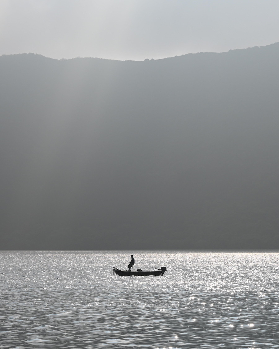 Silhouette of a person standing in a small boat on a shimmering lake, overlooked by misty mountains. 