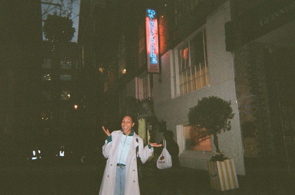 A person smiles broadly, standing outside at night underneath a neon sign that says Pineapple Studios. 