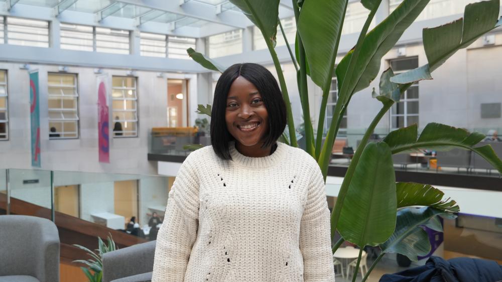 A Black woman with shoulder-length dark hair and a white jumper is smiling at the camera. Behind her there is plant with big, green leaves and an atrium.