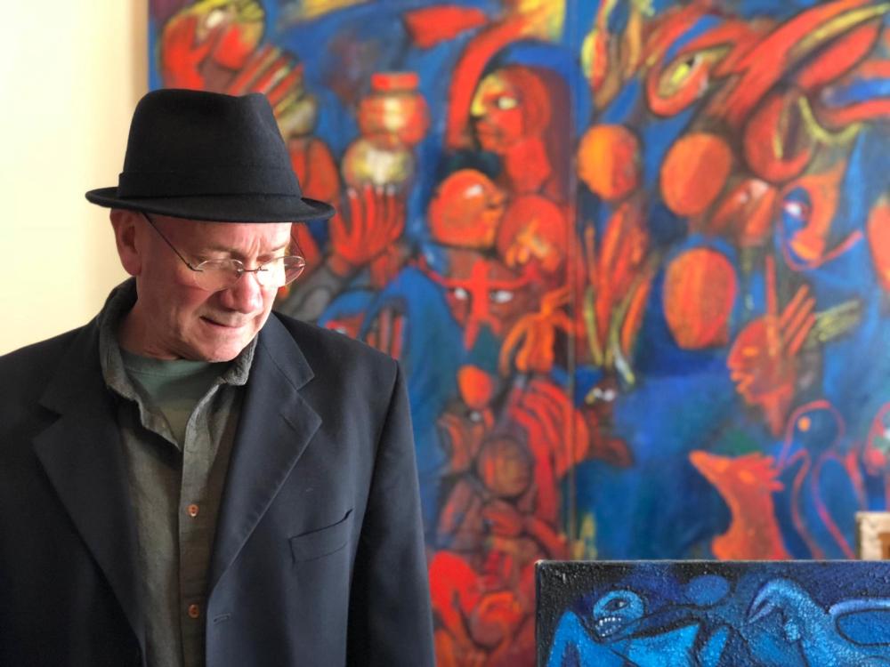 A person wearing a black fedora and dark blazer stands indoors in front of a large, vibrant abstract painting filled with red, blue, and orange figures. A smaller artwork with bold blue shapes is visible in the lower right foreground.