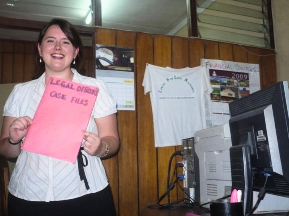 A woman standing in an office holding a pink folder labeled ‘Legal Officers Case Files.’ The office has wood-paneled walls, a desk with a computer, calendars, and a T‑shirt pinned to the wall in the background.
