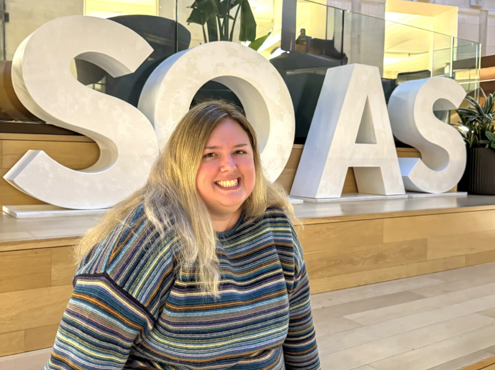 A woman with long, light-blonde hair wearing a striped sweater, seated indoors in front of large white letters spelling ‘SOAS’ in a modern building with wooden steps and glass railings.