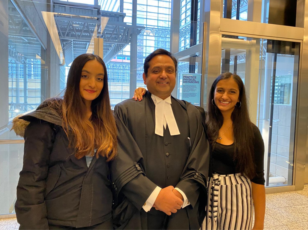 Three people standing together indoors in front of large glass windows and an elevator. The person in the centre is wearing formal barrister robes, while the two people on either side are dressed in modern clothing and smiling toward the camera.