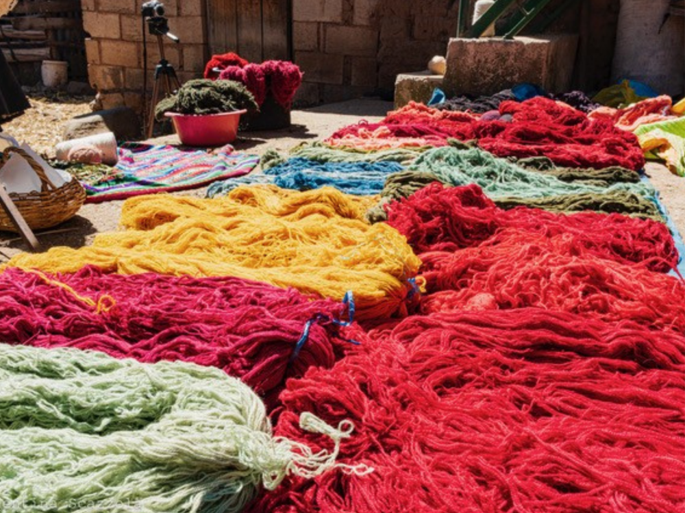 Brightly coloured bundles of yarn in red, yellow, green, and blue spread out on the ground to dry in the sun, surrounded by baskets, bowls, and tools in an outdoor workshop setting.