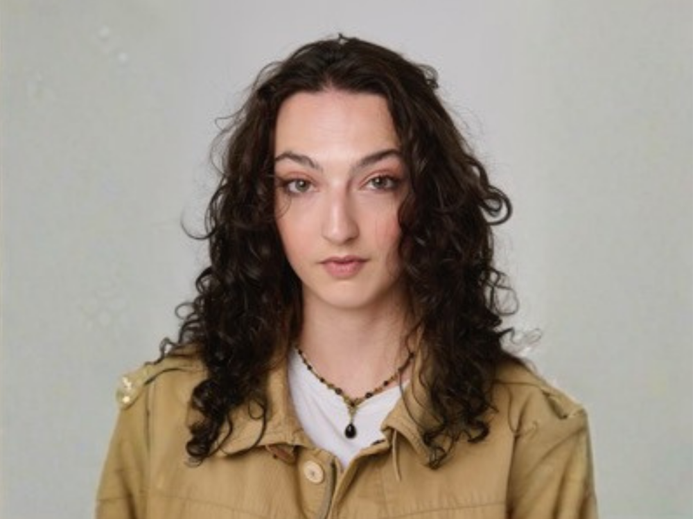 A woman with long, dark curly hair wearing a beige jacket over a white top and a beaded necklace, posed against a light grey background