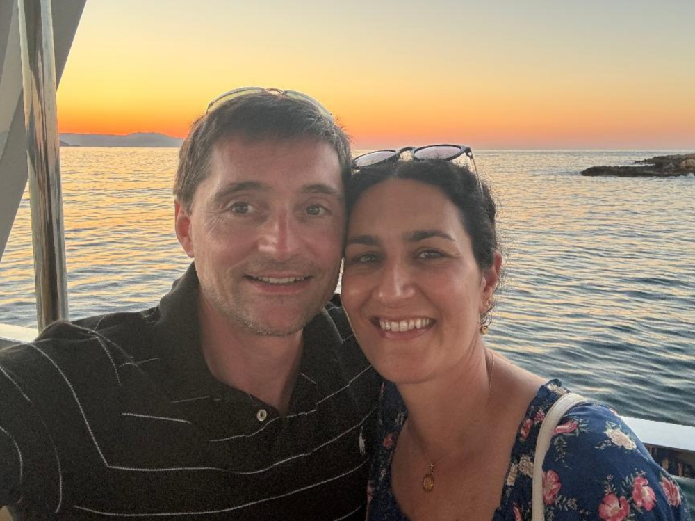 Two people posing together in front of a calm sea at sunset, with orange and pink tones in the sky and a rocky shoreline in the background.