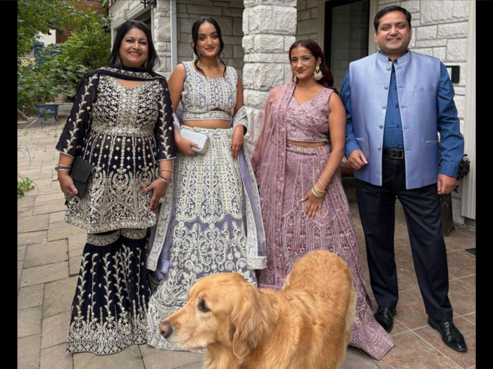 A group of four people dressed in ornate traditional South Asian clothing standing outside a house, with a golden retriever in the foreground.