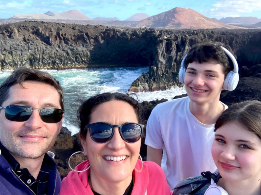 Four people taking a group photo at a rocky coastal viewpoint, with waves crashing against dark volcanic cliffs and distant mountains under a blue sky.