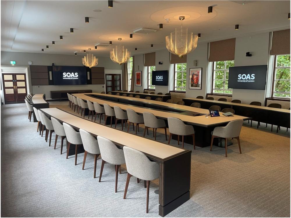 Large conference-type room with rows of tables and chairs, three screens displaying ‘SOAS University of London'.