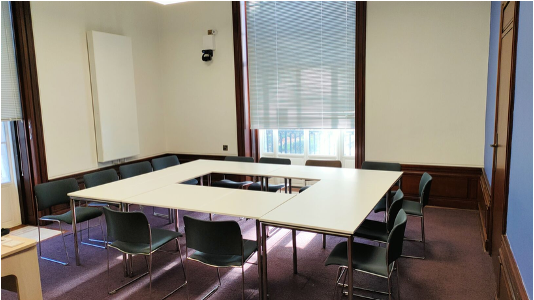 Ten chairs around tables arranged in a rectangle, with two high windows and blinds drawn.