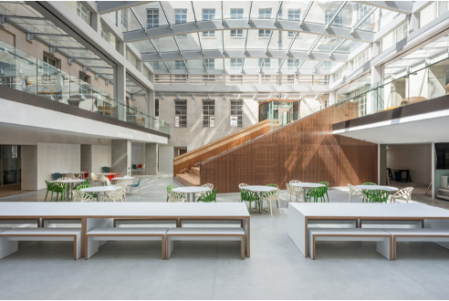 Indoor atrium with a glass ceiling and natural light; white benches in the foreground, circular tables with green and white chairs in the middle, and a wooden staircase in the background