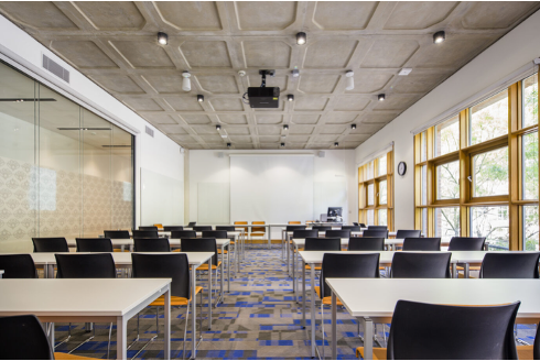 Two rows of white desks and black chairs, glass screen on left, large windows on right, and projector screen at the front.