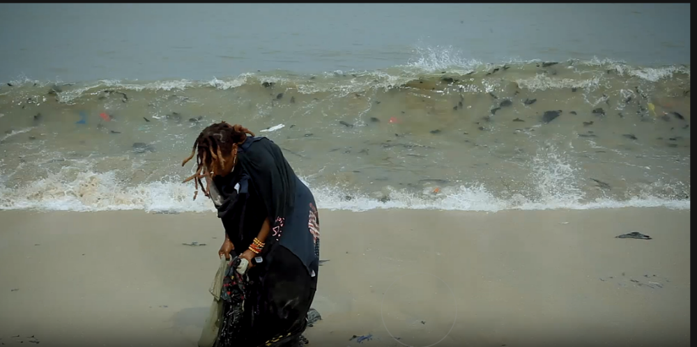 Person collecting litter on a sandy beach, with debris visible in the water behind them.