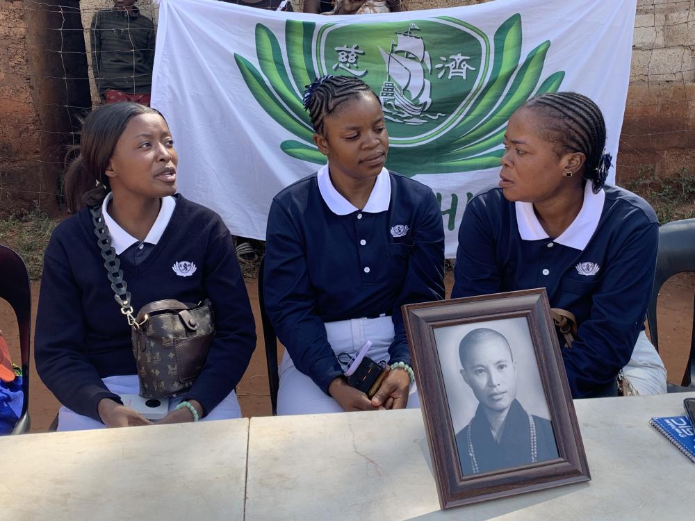 three female volunteers in Tzu Chi uniform, at a table with Cheng Yen's portrait, and the flag of Tzu Chi in the background