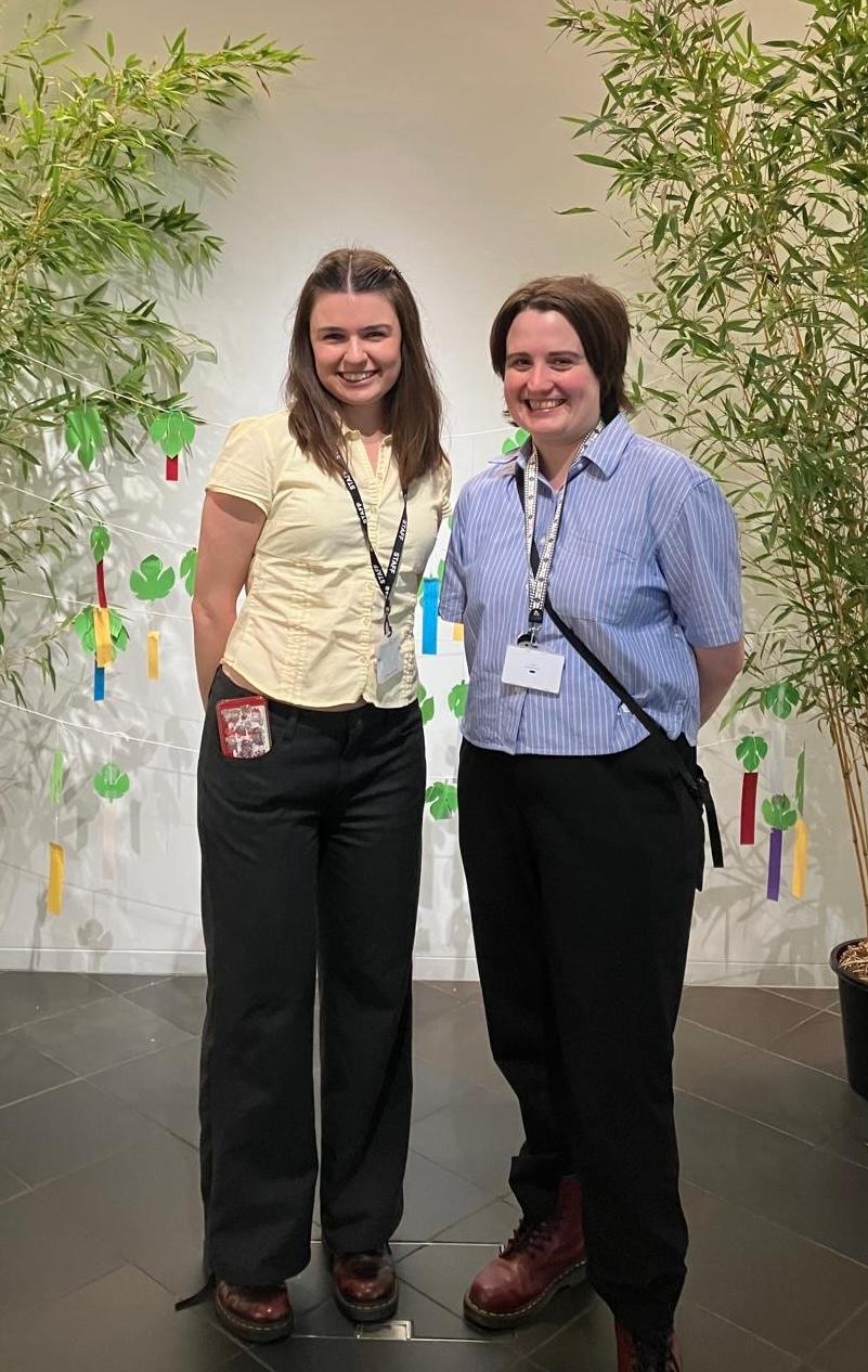 Two smiling women wearing event lanyards pose indoors between bamboo plants and colourful hanging decorations.