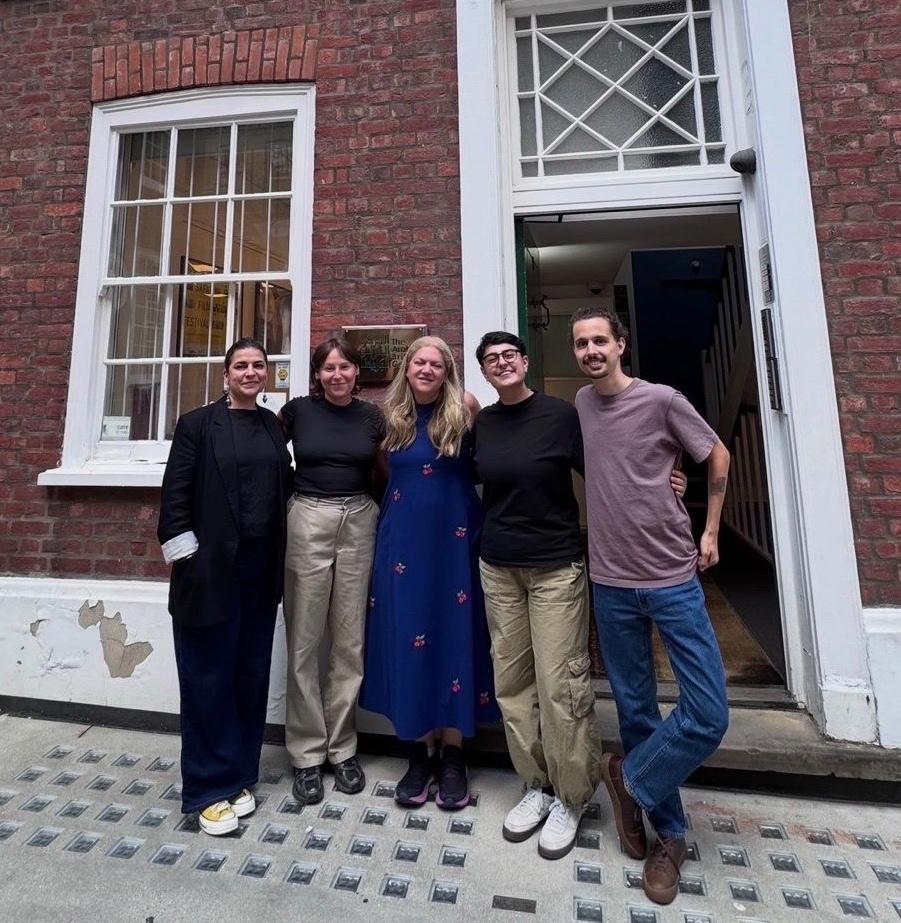 Five smiling adults pose outside a red-brick building, beside a white-framed window and open doorway