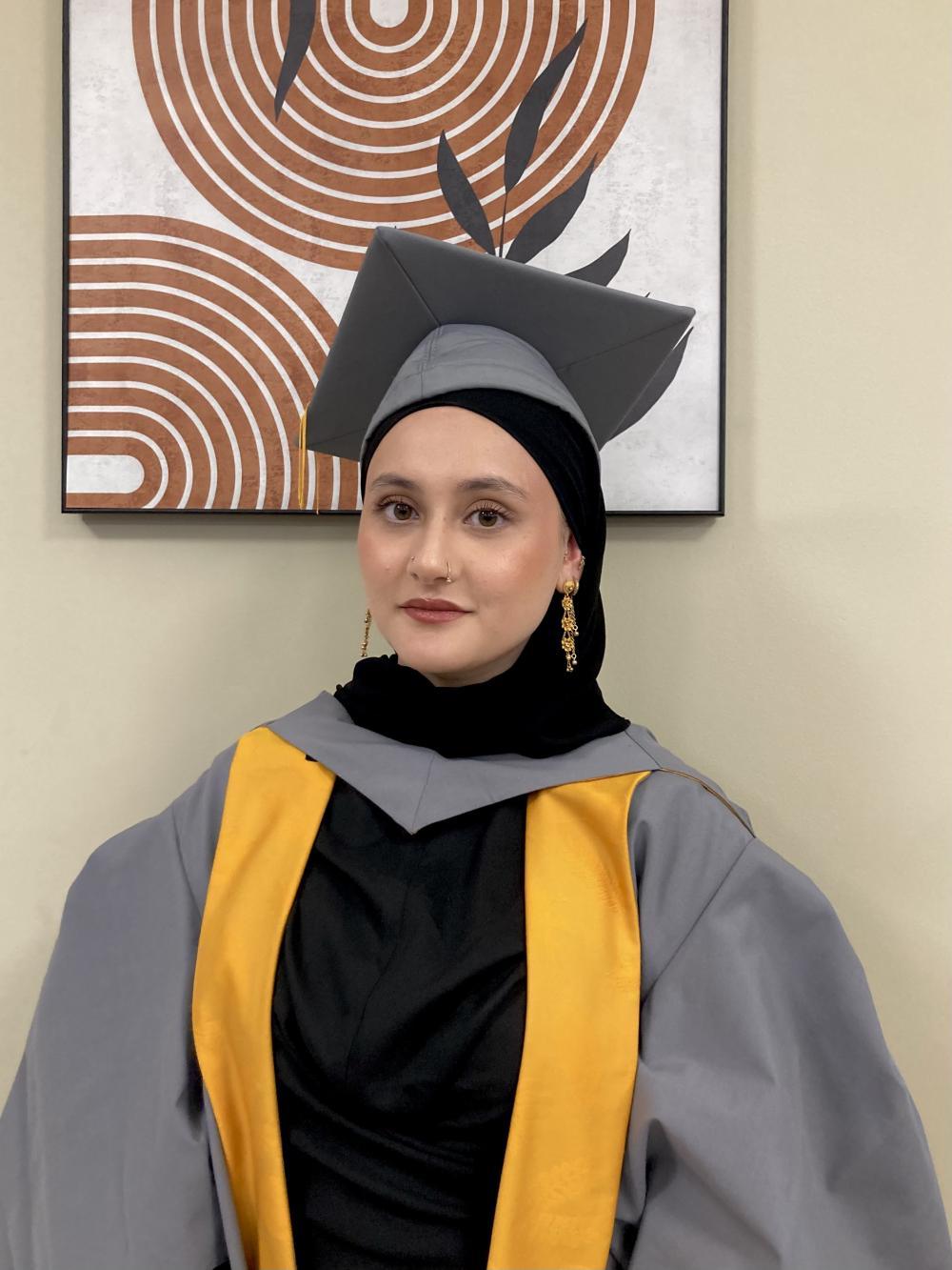 A graduate wearing a grey and yellow gown stands in front of a painting 