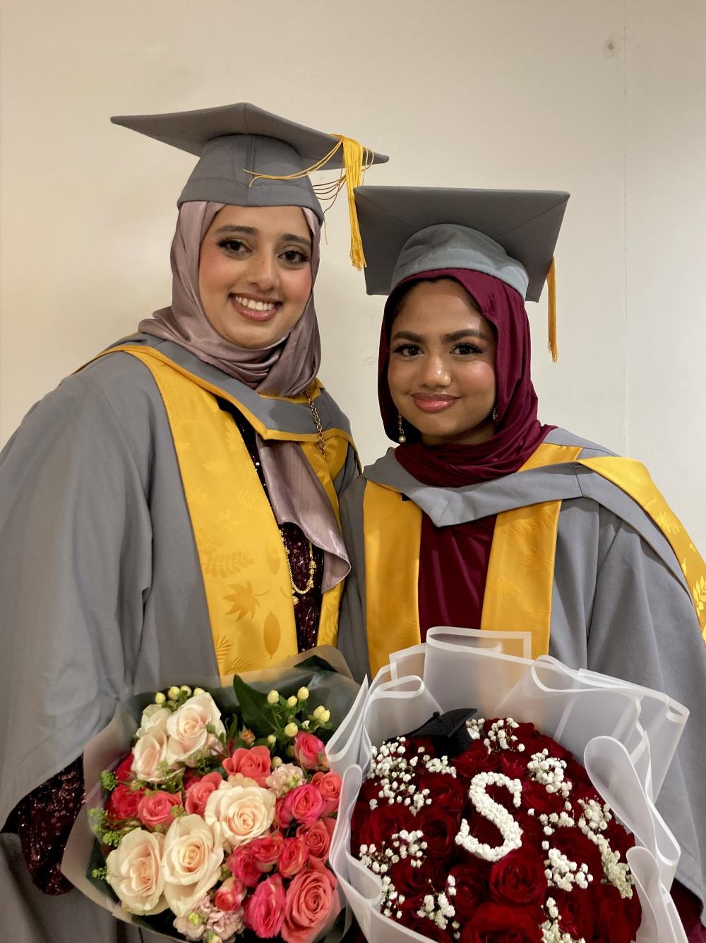 Two graduates in yellow and grey gowns, holding flowers