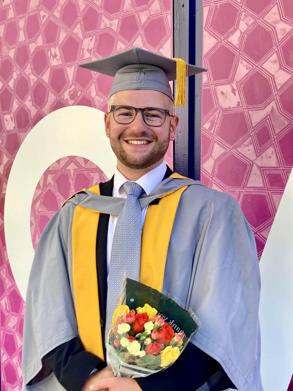 A smiling graduate in grey and yellow gown smiles and holds flowers